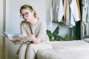 A woman with glasses reading a book while sitting on a bed in a stylish, well-lit bedroom.